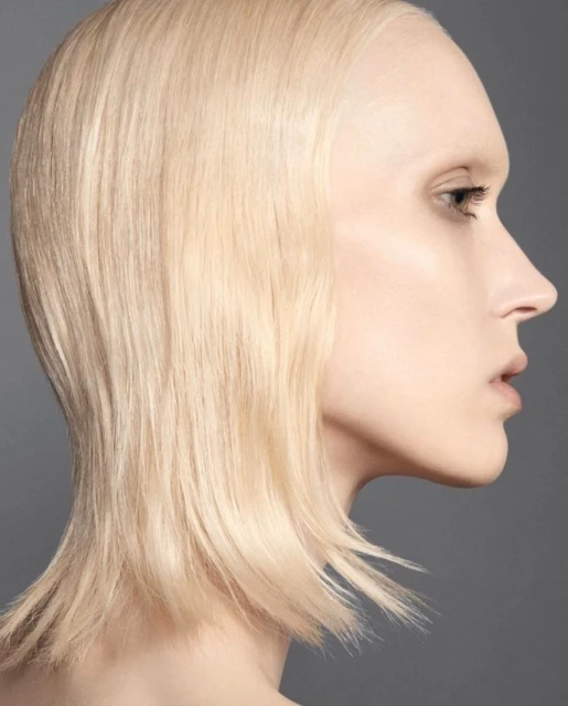 Profile portrait of a young adult woman with short, light blonde hair and fair skin, set against a solid gray background.