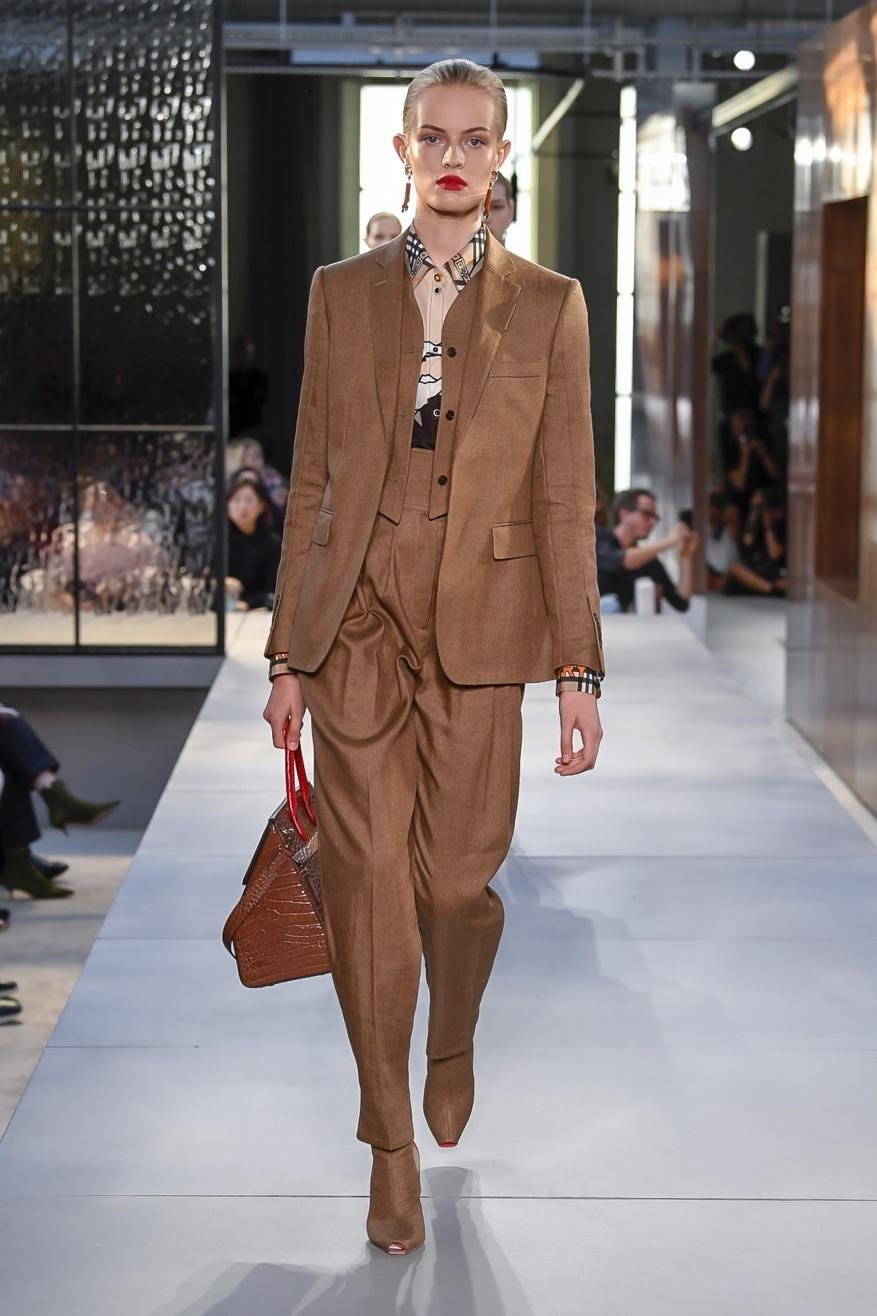 A runway fashion show features a model in a brown suit carrying a red purse, walking on a light grey runway in a well-lit indoor setting.