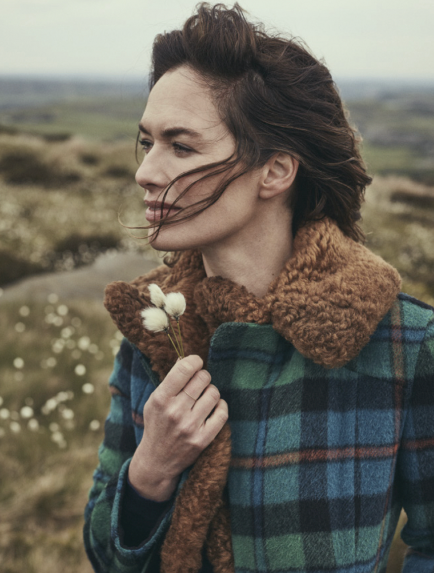 Portrait of woman in plaid coat with fur collar in a windswept outdoor setting, holding small white flowers, against a blurred landscape.