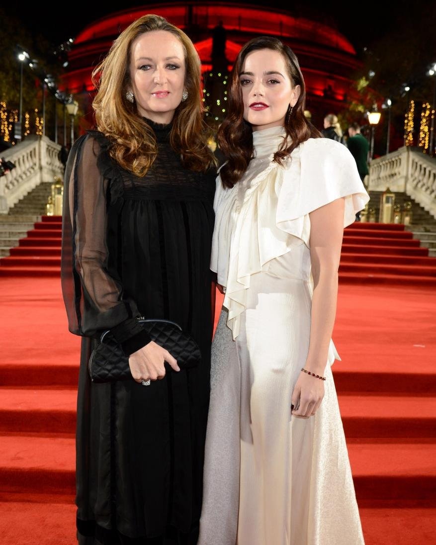 Two women pose on red carpet in evening gowns, one black and one white, at an event with a warmly lit architectural backdrop.