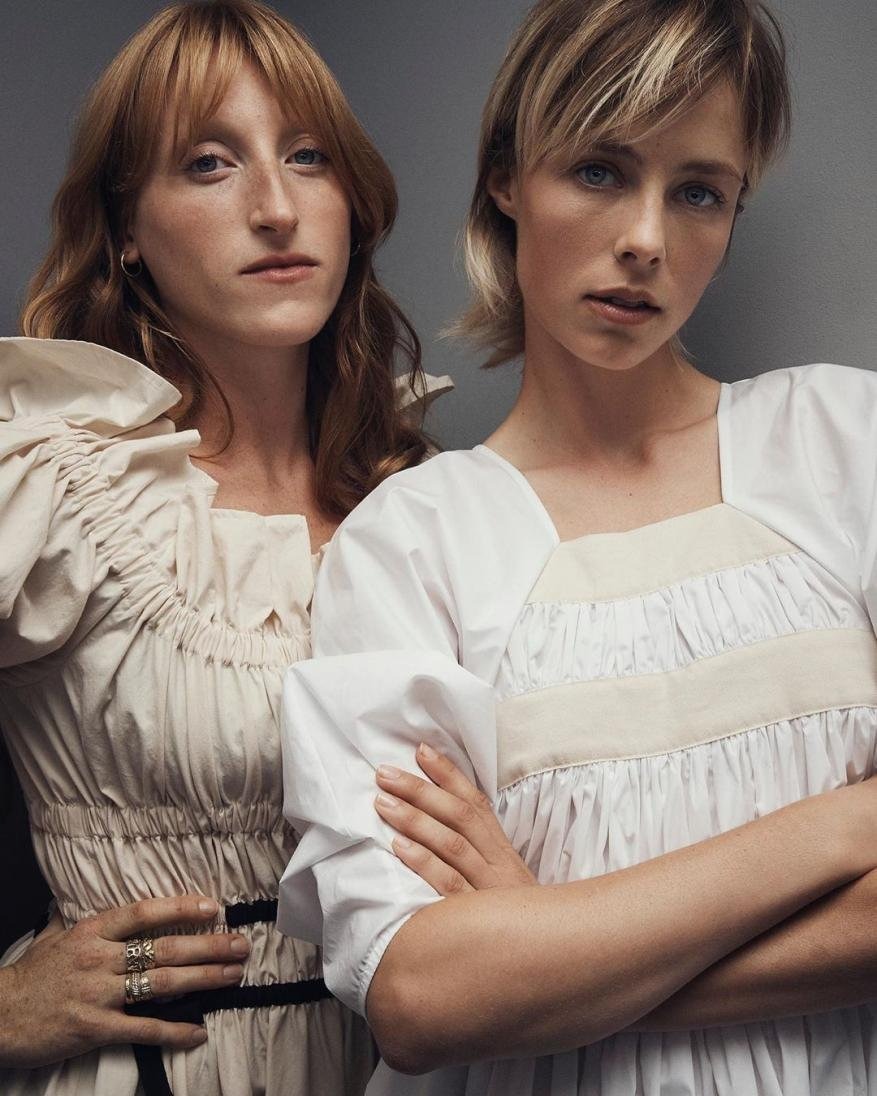 Two adult females in white dresses pose indoors in a medium shot against a gray background, showcasing a fashion editorial style.