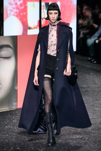 Runway fashion show featuring a stylish adult woman wearing a navy blue cape coat, floral top, shorts, lace-up boots, and a purse.