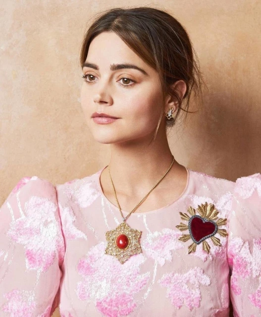 Portrait of an adult female with fair skin wearing a pink floral brocade dress, gold necklace, and heart-shaped brooch against a tan background.