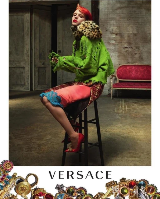 A woman with orange hair sits on a stool wearing a green sweater, pink slip dress, and red shoes in an indoor studio environment.