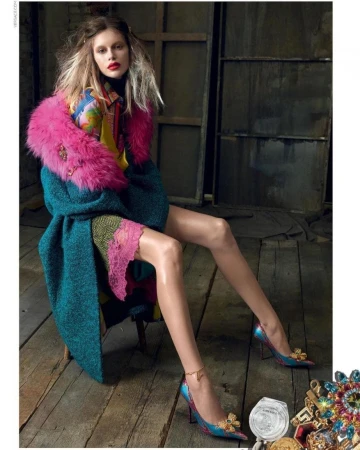 Fashion shot of a woman sitting on a chair, wearing a turquoise coat with a pink fur collar and patterned heels on a wood floor.