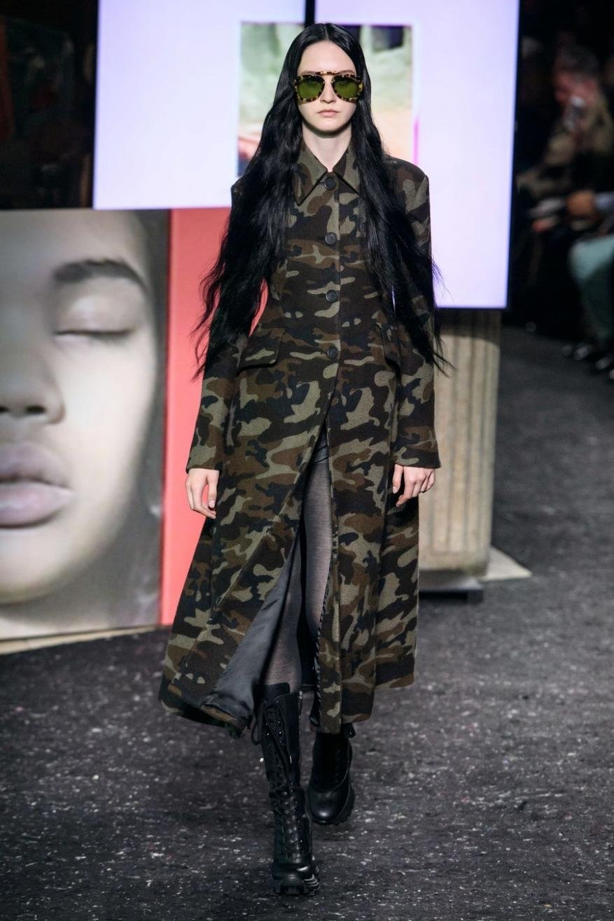 Runway fashion show featuring a female model in a camouflage coat and sunglasses, walking on a dark runway with stage lighting.