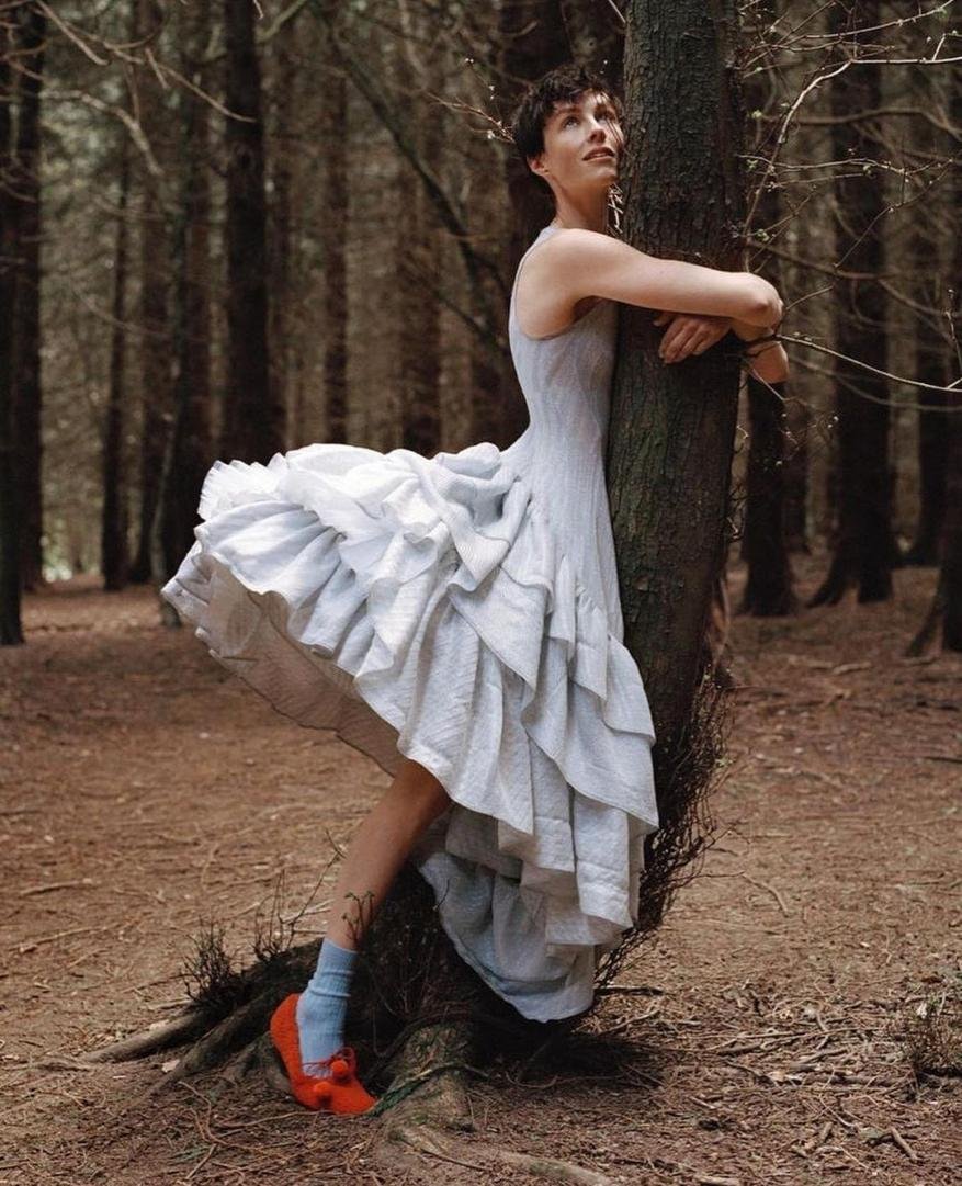 Woman in pale blue dress hugging tree trunk in forest, wearing light blue socks and red shoes