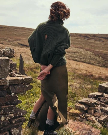Woman seen from behind wearing an olive green turtleneck sweater and long skirt standing outdoors near a stone wall.