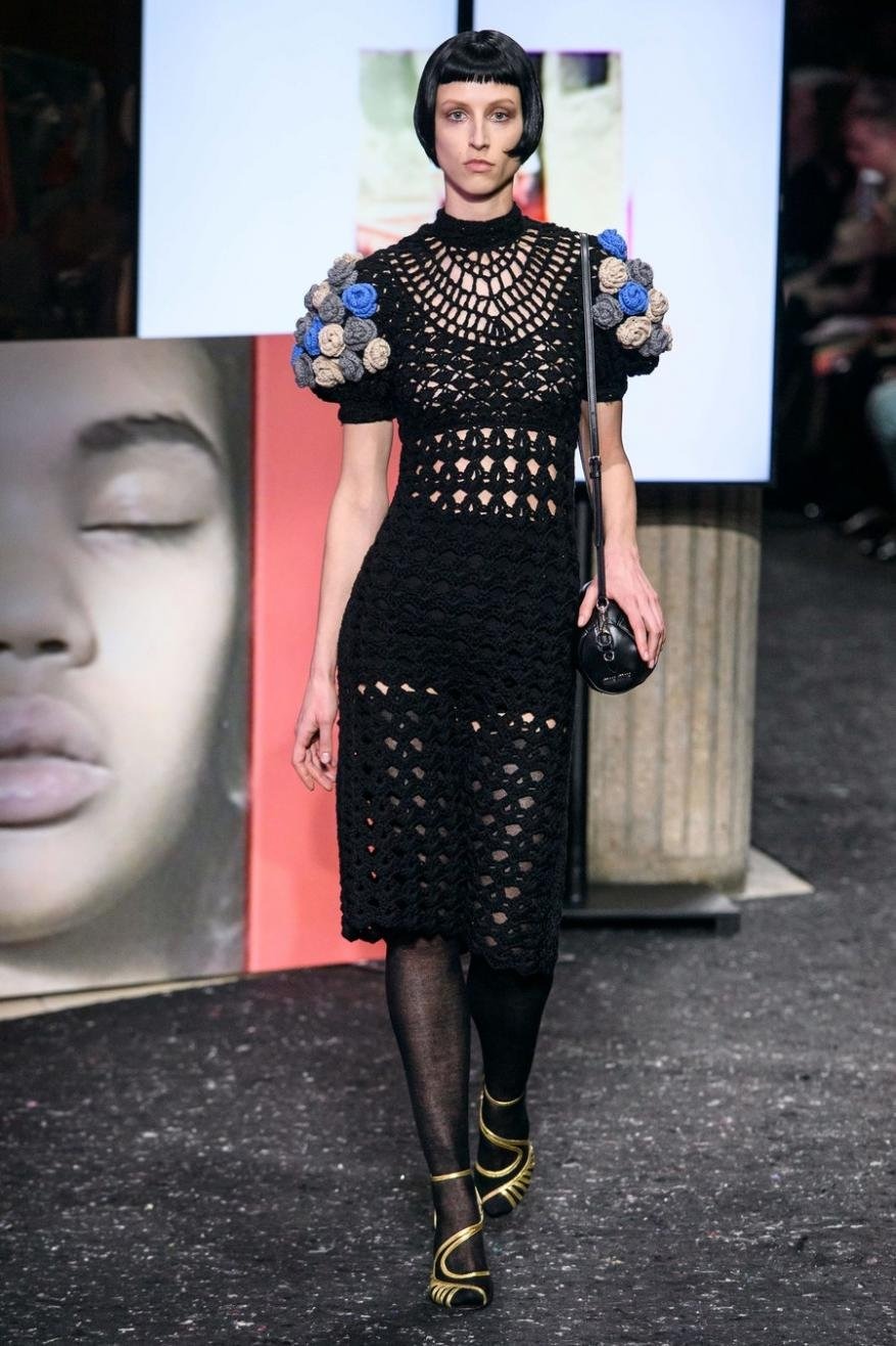 Runway shot of a fair-skinned fashion model in a black crochet dress with blue floral sleeves, black tights, and gold strappy shoes.