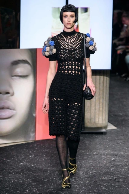 Runway shot of a fair-skinned fashion model in a black crochet dress with blue floral sleeves, black tights, and gold strappy shoes.
