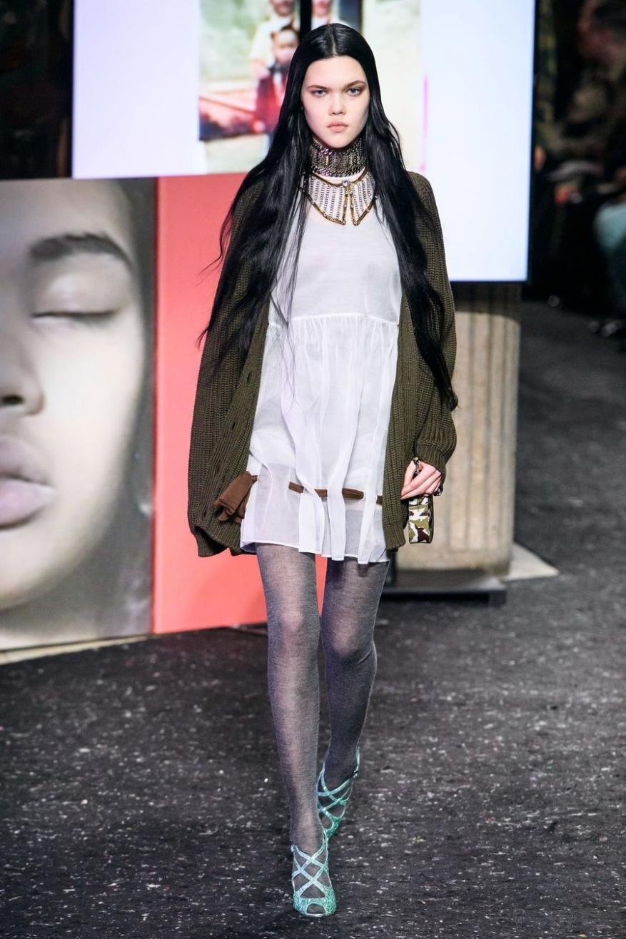 A fashion model walks the runway in a white dress, green cardigan, sheer tights, and strappy heels during a fashion show event.