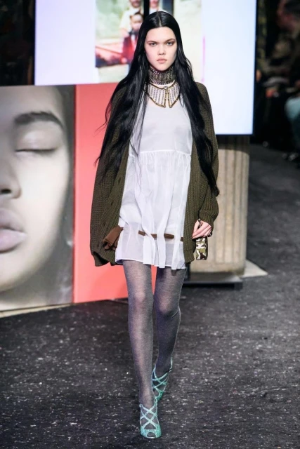 A fashion model walks the runway in a white dress, green cardigan, sheer tights, and strappy heels during a fashion show event.