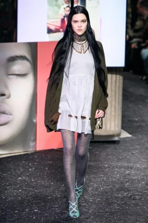 A fashion model walks the runway in a white dress, green cardigan, sheer tights, and strappy heels during a fashion show event.