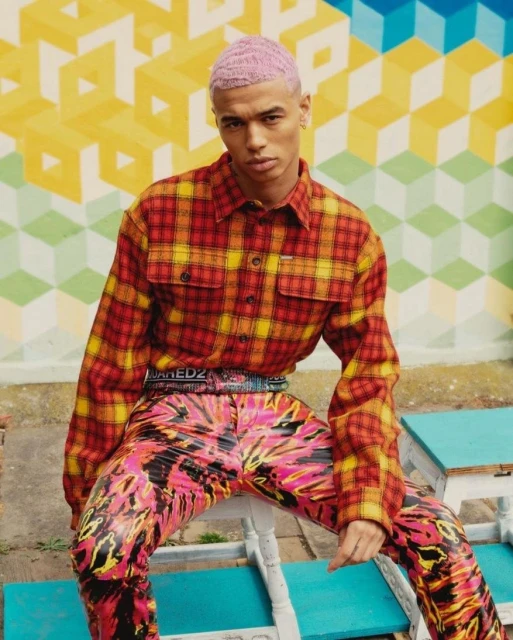 Young adult male model with pink hair poses while sitting outside in red and yellow plaid shirt and brightly colored pants.