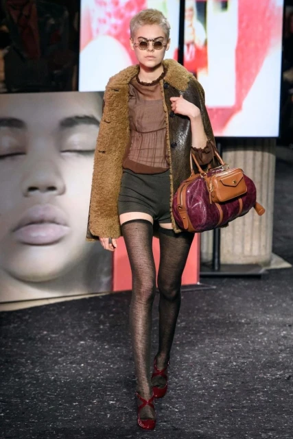 A woman on a runway presents fashion, wearing a shearling coat, dark shorts, patterned stockings, and heeled shoes, holding a bag.