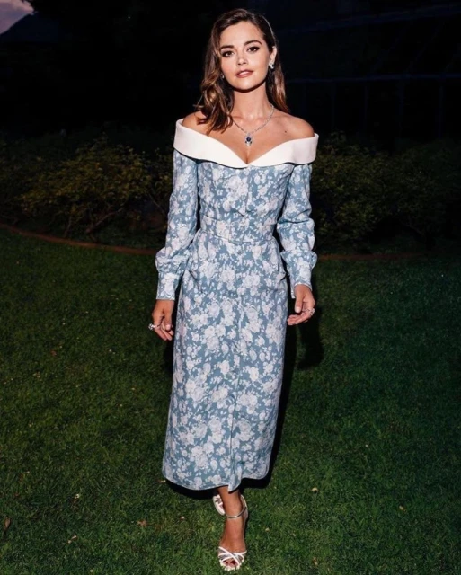 A woman in a long-sleeved blue floral dress stands on green grass, accessorized with jewelry and strappy heels, at eye level.