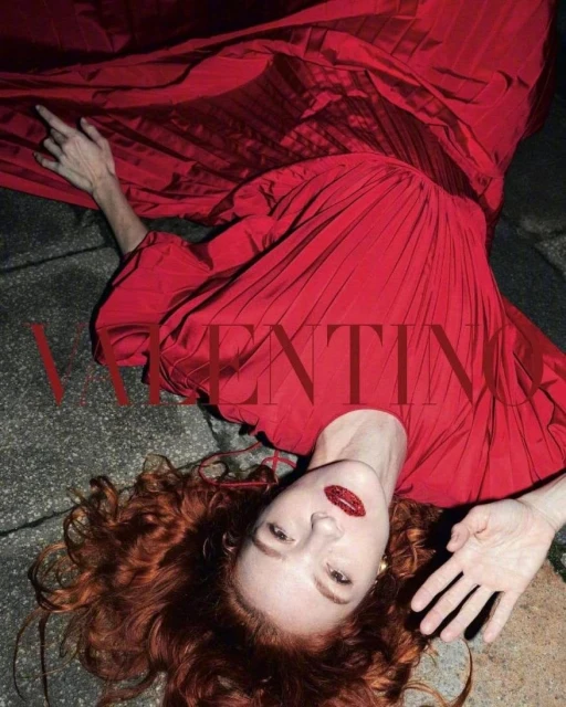 Woman lies on grey surface in a red Valentino gown. Overhead angle, looking at the camera. Red hair and lipstick.