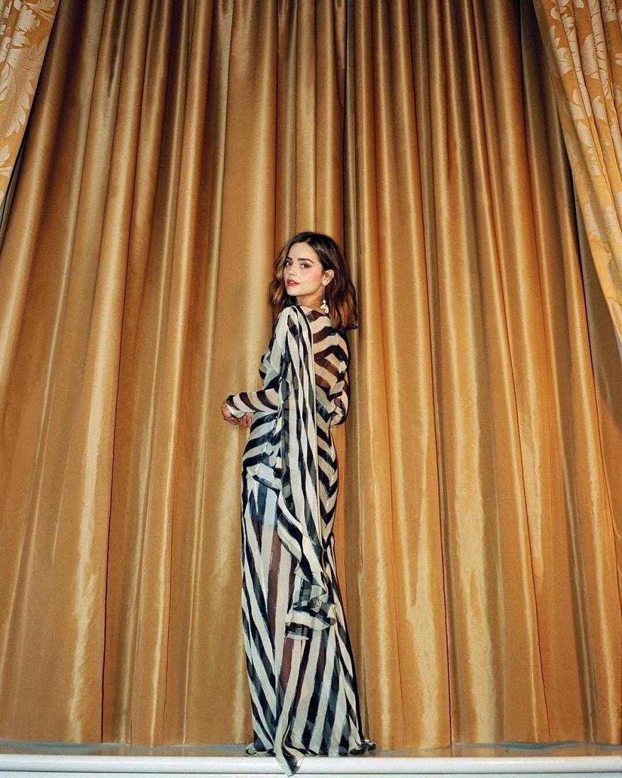 A woman in a striped dress stands in front of golden drapes in a studio setting, posing and glancing over her shoulder.