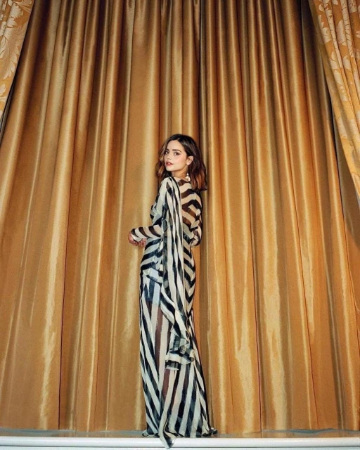 A woman in a striped dress stands in front of golden drapes in a studio setting, posing and glancing over her shoulder.
