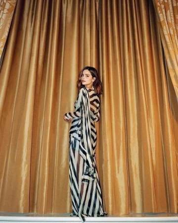 A woman in a striped dress stands in front of golden drapes in a studio setting, posing and glancing over her shoulder.