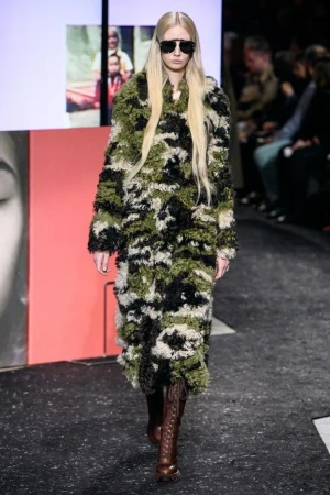 Female fashion model walks runway wearing a camouflage coat and sunglasses in a fashion show with audience in the background.