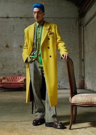 Man with vibrant blue hair wearing a bright yellow overcoat, patterned green shirt, and plaid trousers, posing in a loft.