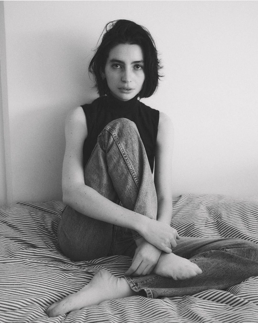 Black and white portrait of a young woman with dark hair sitting on a striped bed, wearing jeans and a sleeveless top, looking at camera.