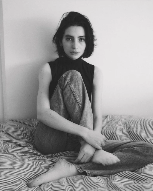 Black and white portrait of a young woman with dark hair sitting on a striped bed, wearing jeans and a sleeveless top, looking at camera.