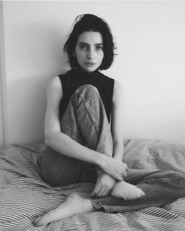 Black and white portrait of a young woman with dark hair sitting on a striped bed, wearing jeans and a sleeveless top, looking at camera.