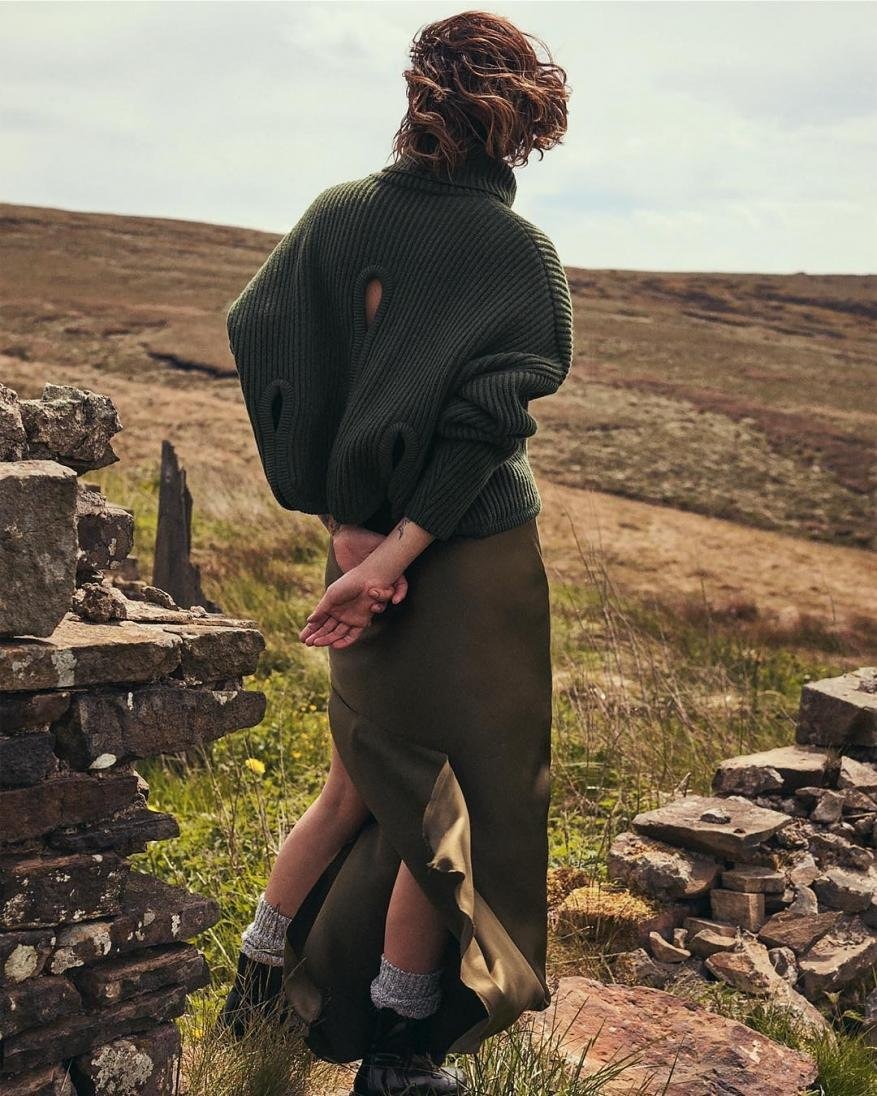 Woman with curly brown hair seen from behind wearing an olive green turtleneck sweater and long skirt standing outdoors on a moor.