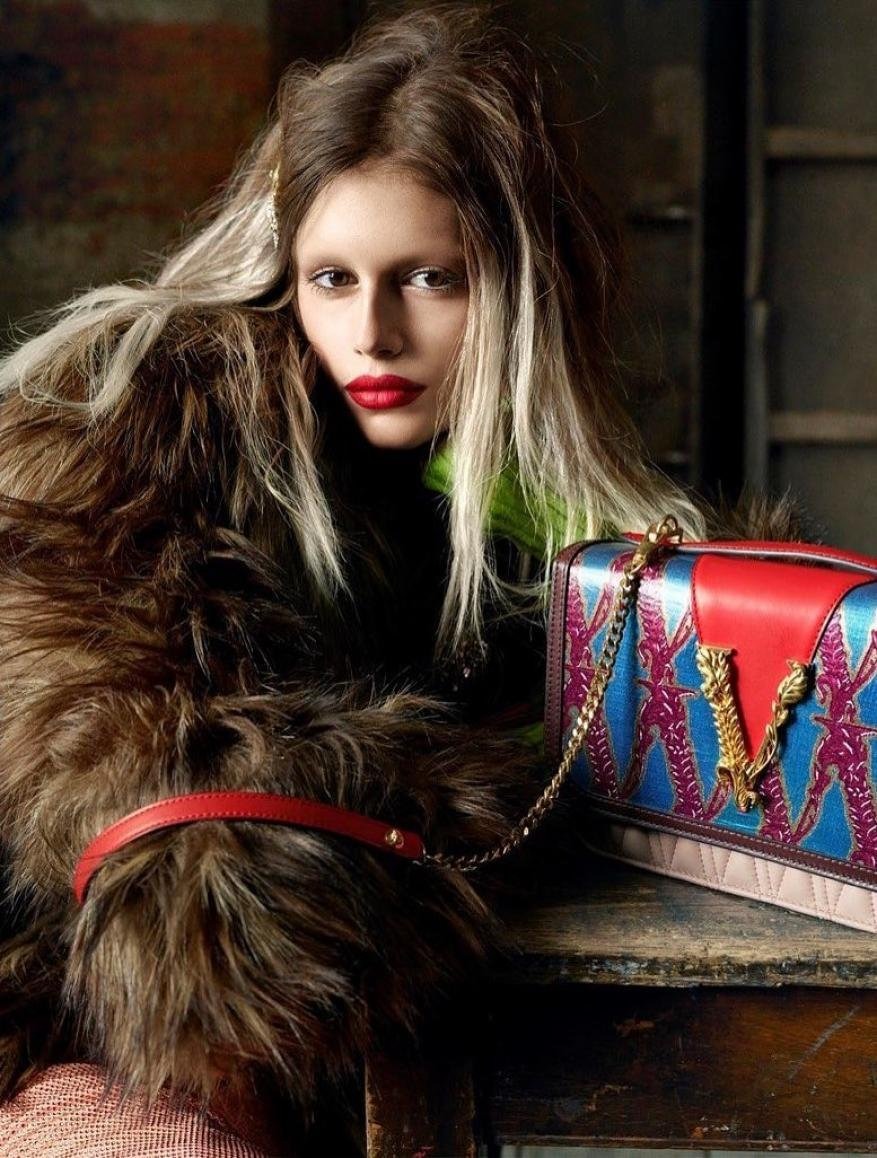 Fashion shot: Woman poses in a fur coat with blond-streaked hair next to a patterned handbag on a distressed table indoors.