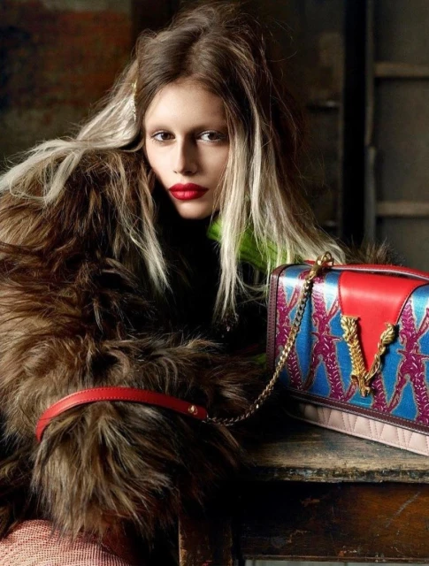 Fashion shot: Woman poses in a fur coat with blond-streaked hair next to a patterned handbag on a distressed table indoors.