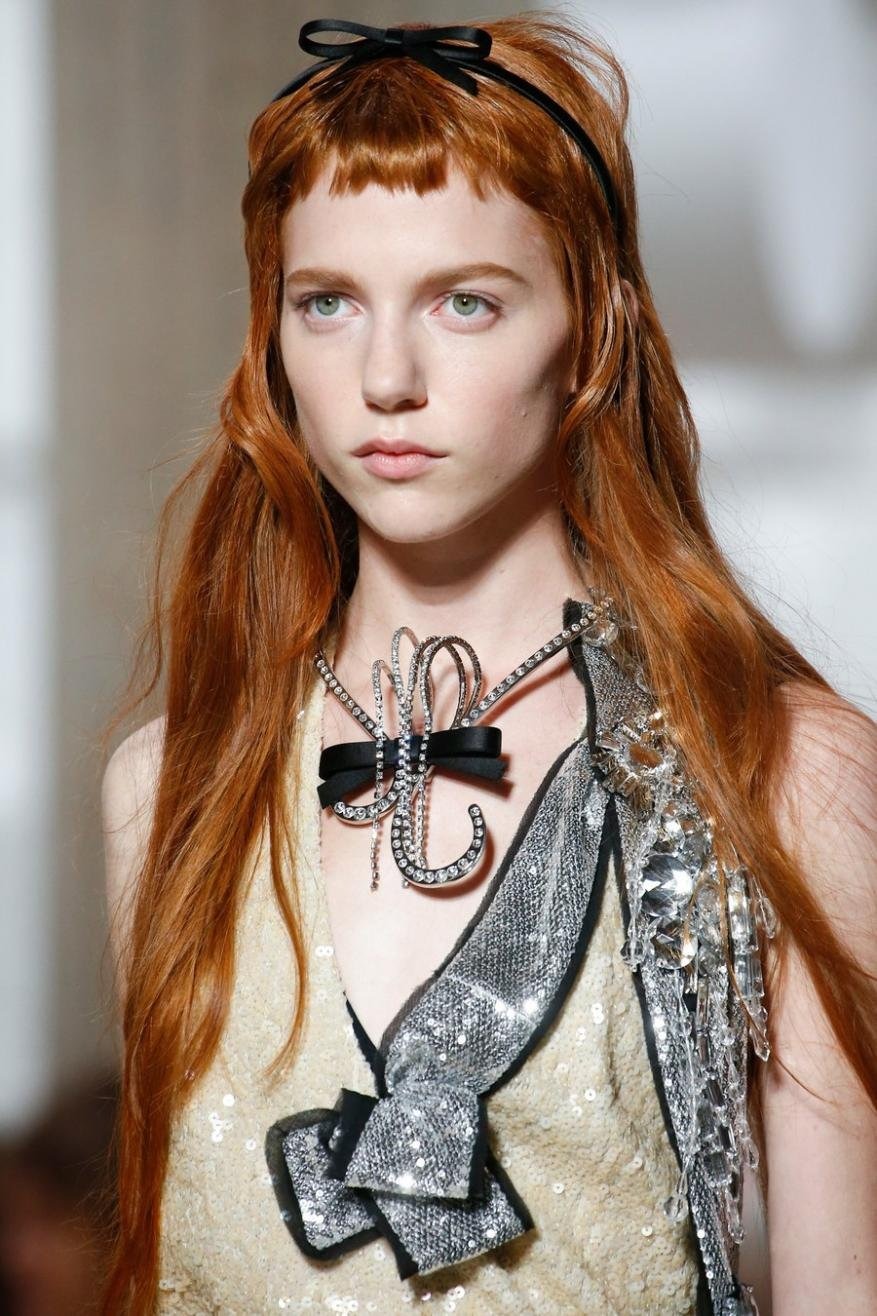 Close-up of a redhead woman wearing an embellished top, necklace, and hair bow on a white background in an indoor setting.