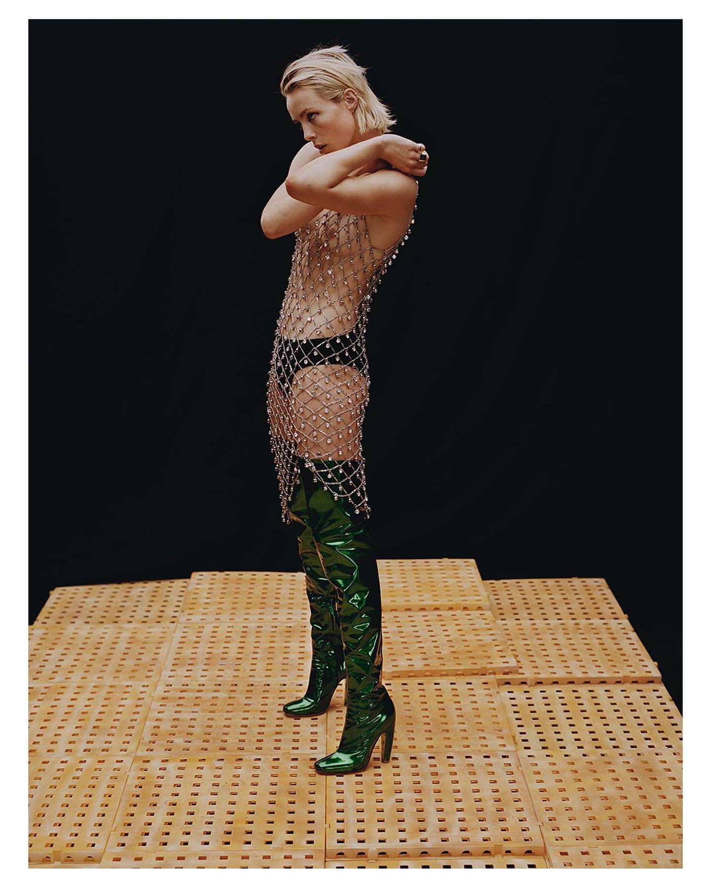 A slender adult woman poses in a translucent beaded dress and green patent-leather thigh-high boots on a lattice platform against black.