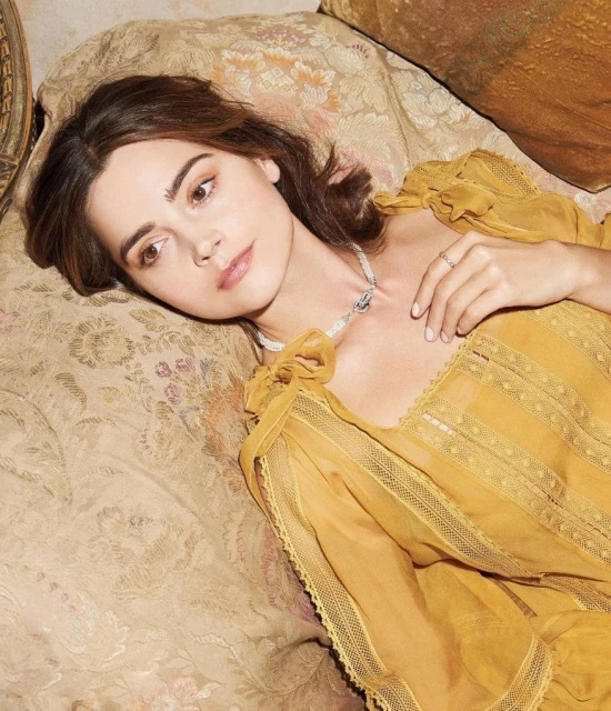 Elegant woman with neutral facial expression lying on a patterned couch wearing a yellow dress and pearl necklace in soft studio light.