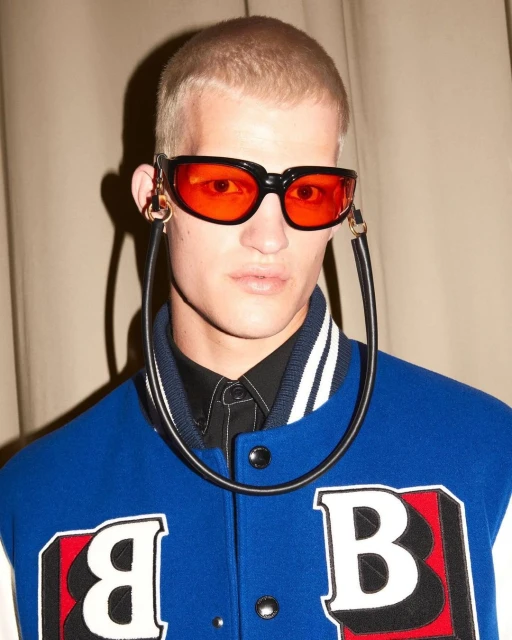 Close-up studio shot of a blonde male model wearing orange sunglasses with black frames and a blue and white letterman jacket.