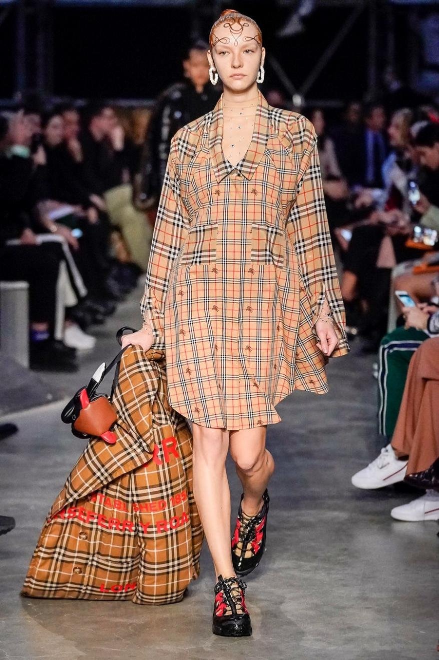 Runway fashion show featuring a model in a tan plaid dress carrying a matching plaid bag, walking on a gray runway.