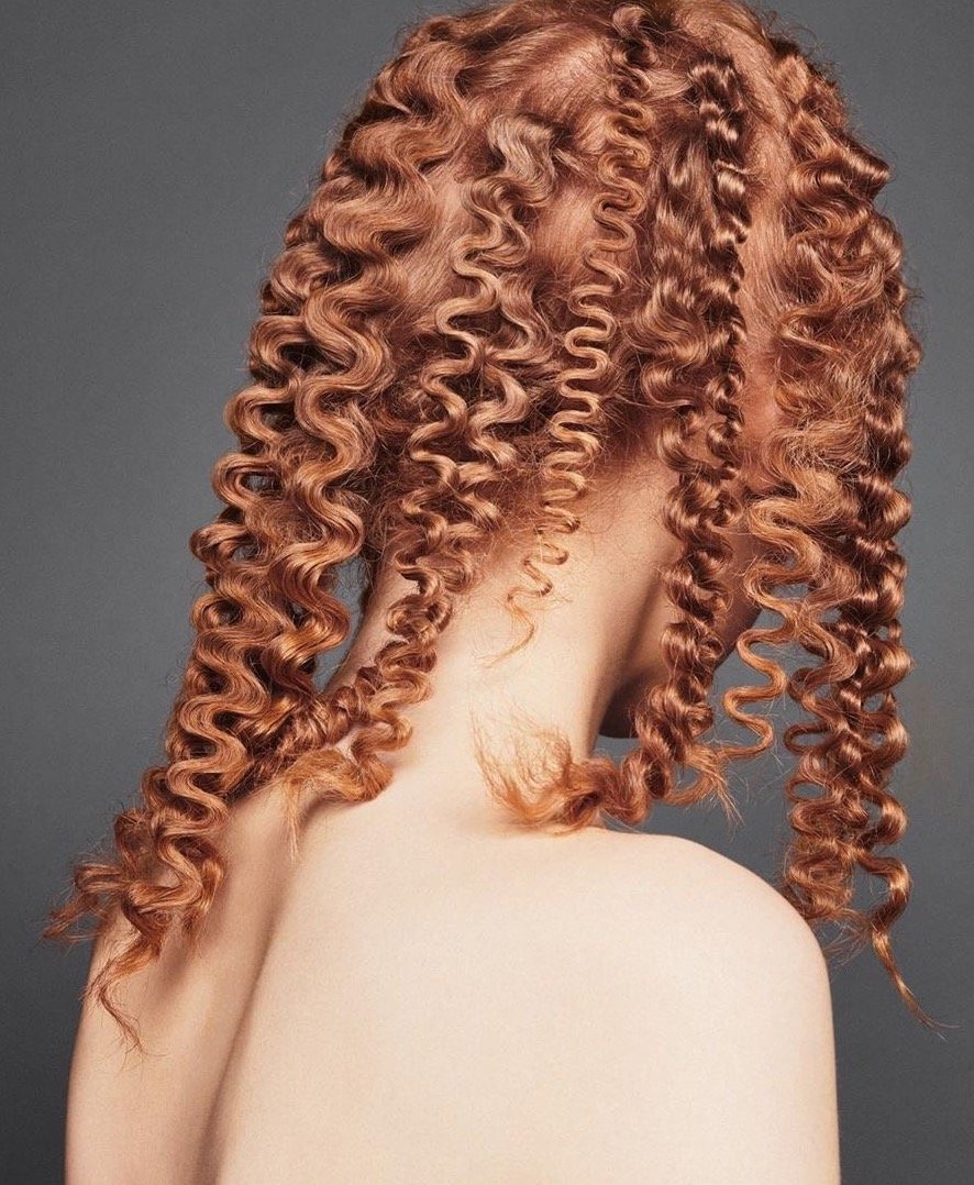Close-up of a fair-skinned woman with shoulder-length, reddish-blonde curly hair against a grey background.