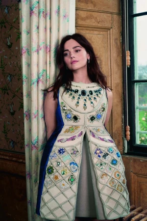 Woman in an elaborate cream and blue dress with jewelled embellishments, standing indoors by a window with floral curtains.