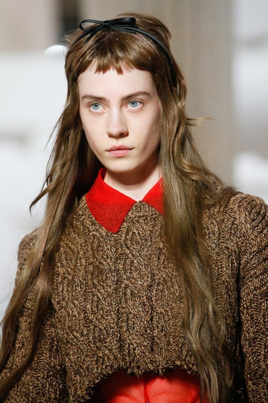 Close-up of a fair-skinned adult female wearing a brown sweater and red collared shirt with a black headband on a bright background.