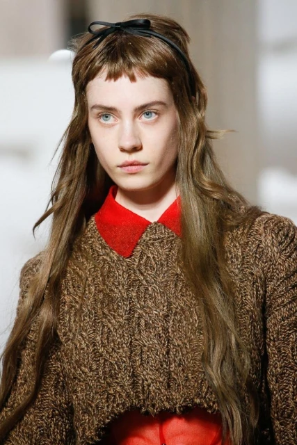 Close-up of a fair-skinned adult female wearing a brown sweater and red collared shirt with a black headband on a bright background.