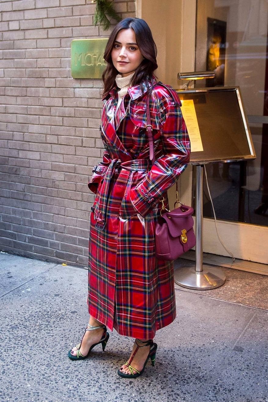 A woman in a red plaid trench coat stands outside a building with a matching purse, showcasing a sophisticated, fashionable outfit.