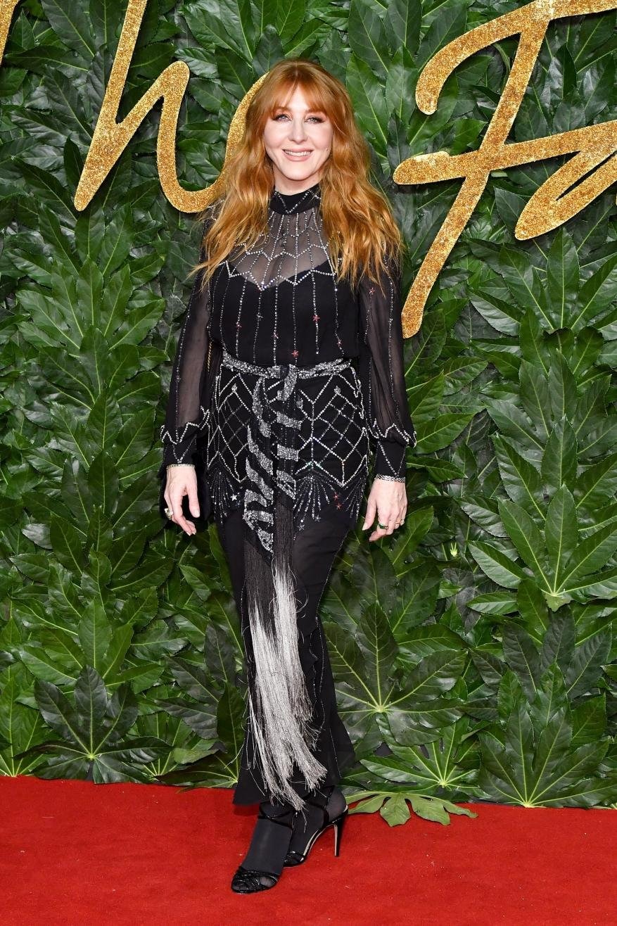 Woman with red hair in a long black dress posing in front of a leafy backdrop at a fashion event, standing on a red carpet.
