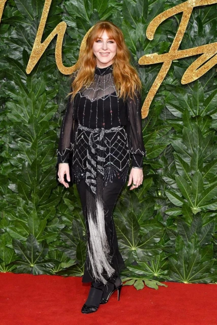 Woman with red hair in a long black dress posing in front of a leafy backdrop at a fashion event, standing on a red carpet.