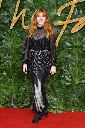 Woman with red hair in a long black dress posing in front of a leafy backdrop at a fashion event, standing on a red carpet.