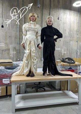 Two women wearing dramatic floor-length dresses standing on a workbench in a concrete warehouse space.