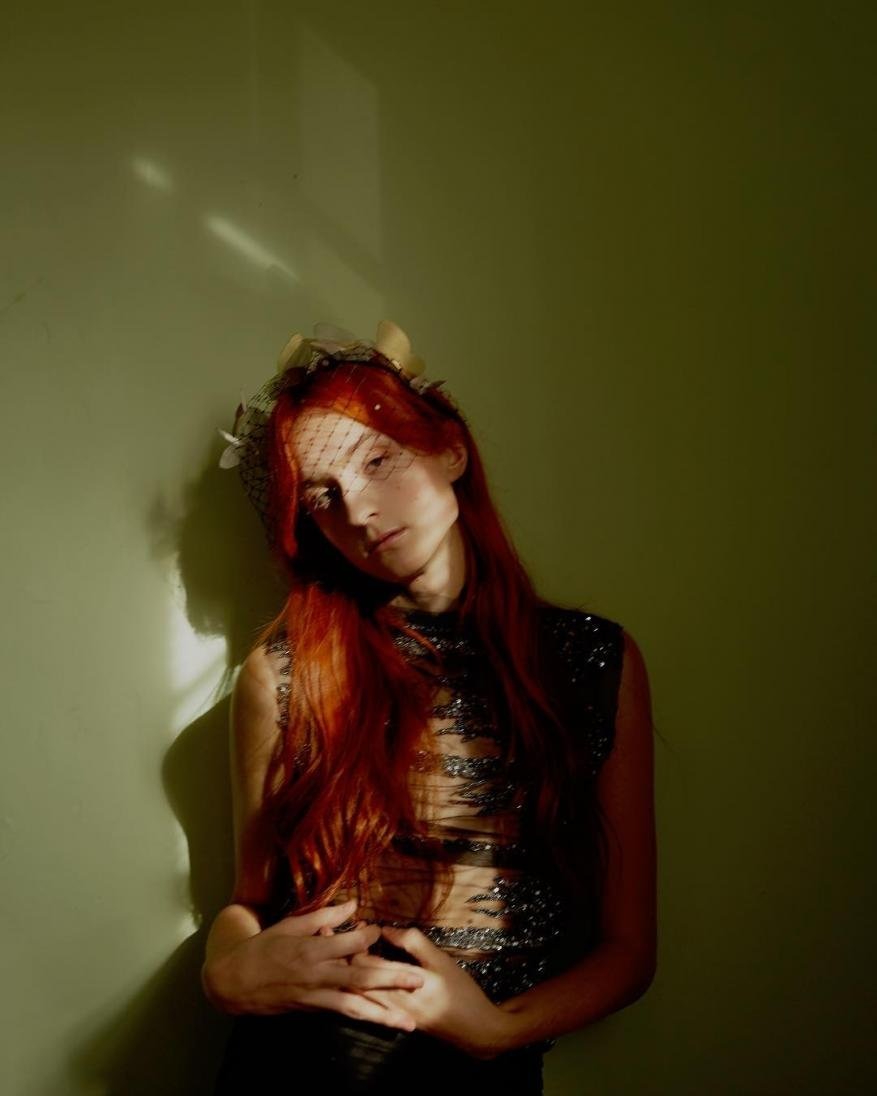 Red-haired woman in a dark top and netting stands in front of a muted green wall with light shining on her, creating shadows.
