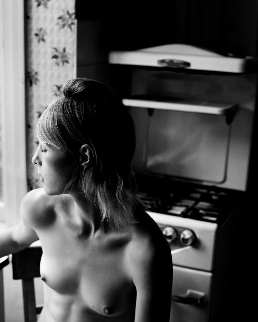 Black and white photo of a topless, fair-skinned woman sitting indoors with a stove and patterned wallpaper in the background.