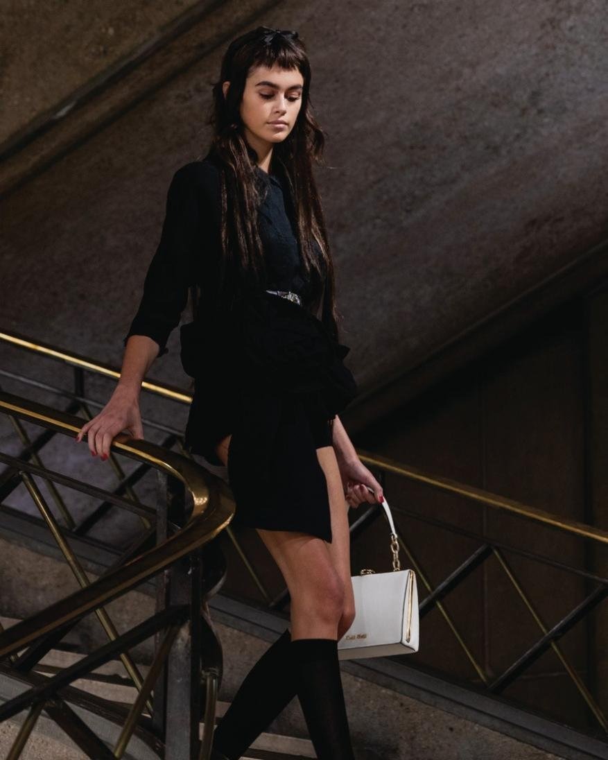 A brunette model walks down a staircase wearing a black mini dress, knee high socks, and carrying a white leather purse.