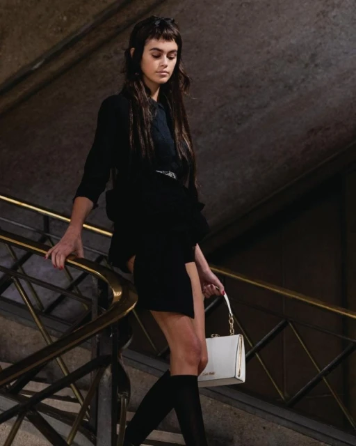 A brunette model walks down a staircase wearing a black mini dress, knee high socks, and carrying a white leather purse.